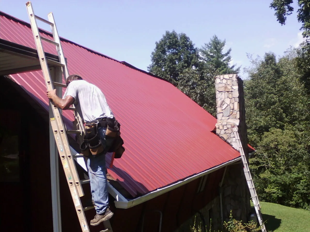 Roofer on a ladder beside a red metal roof and stone chimney — Soffit & Fascia Repair by Roofing Brothers in Irwin